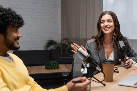 excited brunette podcaster holding professional microphone and talking to blurred indian colleague in yellow jumper sitting near takeaway drink on table in radio studio