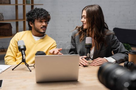 femme brune positive et bouclé homme indien en pull jaune parlant près de microphones, ordinateur portable, boissons à emporter et appareil photo numérique flou dans un studio de radio professionnel