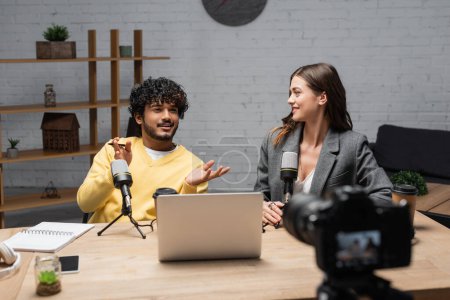 Photo for Positive indian man in yellow jumper gesturing near microphones, laptop, notebook and smartphone while talking to smiling colleague sitting in blazer in front of blurred digital camera in studio - Royalty Free Image