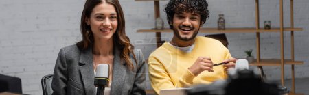 Téléchargez les photos : Femme brune gaie en blazer gris et bouclé homme indien en pull jaune souriant près de microphones professionnels tout en travaillant dans un studio de radio moderne, bannière - en image libre de droit