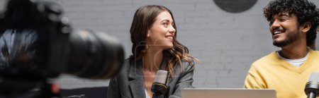 Photo for Bearded indian man in yellow jumper and charming brunette woman in grey blazer smiling at each other while recording podcast in front of studio microphones and blurred digital camera, banner - Royalty Free Image
