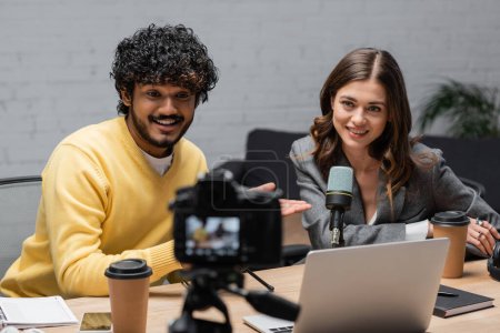 Photo for Cheerful indian man in yellow jumper pointing at happy brunette colleague near laptop, paper cup and notebooks while recording podcast on blurred digital camera in professional radio studio - Royalty Free Image