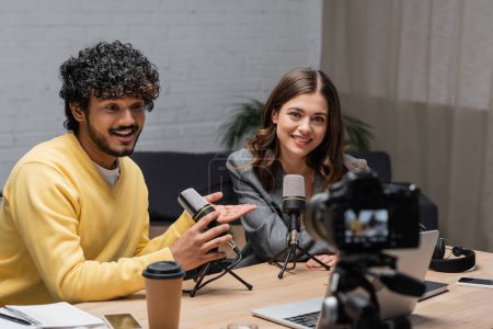 joyful and curly indian man in yellow jumper talking in microphone and pointing at charming colleague near laptop, coffee to go and smartphones in front of blurred digital camera in broadcasting studio