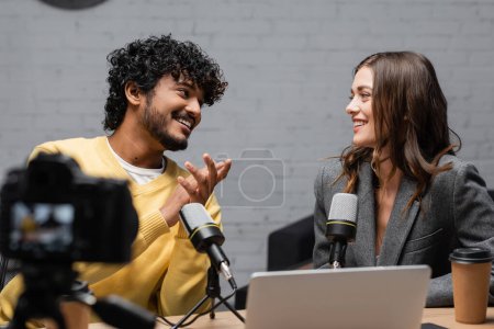 Photo for Curly and bearded indian man in yellow jumper smiling at charming colleague sitting in grey blazer near microphones, laptop and coffee to go in front of blurred digital camera in studio - Royalty Free Image