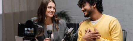 honest and bearded indian man touching chest and talking to charming brunette colleague near microphones and blurred digital camera in radio studio, banner