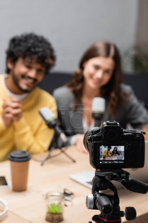 focus on professional digital camera recording blurred and happy interracial podcasters near professional microphones and coffee to go on table in radio studio