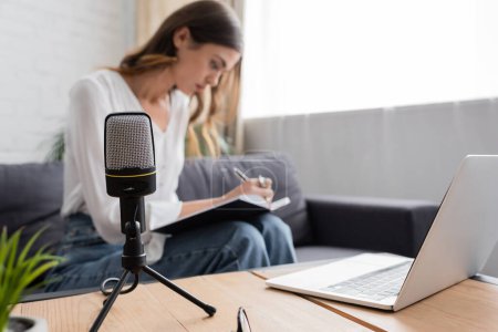 focus on professional microphone and laptop on table near blurred charming broadcaster writing in notebook while sitting on couch in modern studio
