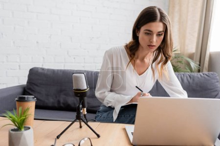 pensive brunette woman sitting on couch and looking at laptop while preparing podcast near professional microphone, takeaway drink and flowerpot on table in radio studio