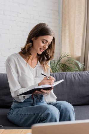 cheerful and pretty broadcaster with brunette hair wearing white blouse and writing in notebook with pen while sitting on couch and preparing podcast in radio studio