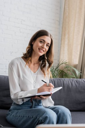 charming and happy brunette woman in white blouse and denim jeans looking at camera while sitting on grey couch and writing in notebook with pen in radio studio