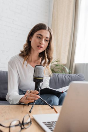 charming and thoughtful brunette woman in white blouse holding professional microphone and notebook while sitting near laptop and blurred eyeglasses in radio studio