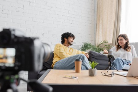hombre indio rizado en jersey amarillo y mujer morena en blusa blanca hablando en el sofá junto al micrófono profesional, portátil, café para llevar y teléfono móvil frente a la cámara digital borrosa