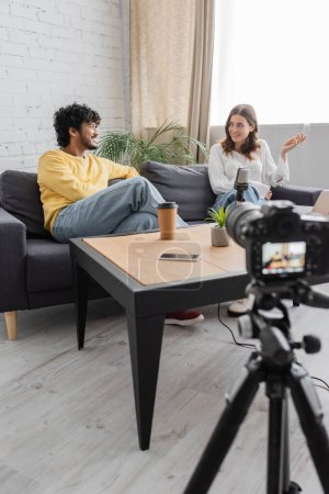 Photo for Smiling interracial couple of podcasters talking on sofa near table with microphone, paper cup, flowerpot and smartphone with blank screen in front of blurred digital camera in radio studio - Royalty Free Image