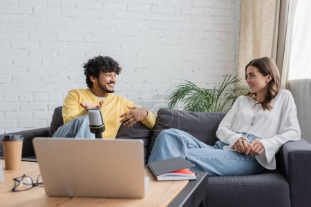 Photo for Young indian man in yellow jumper and smiling brunette woman sitting on couch near laptop, microphone, notebook, paper cup and blurred eyeglasses while talking in modern studio - Royalty Free Image