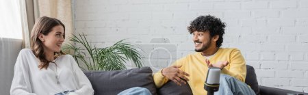hombre indio rizado en jersey amarillo gesto mientras está sentado en el sofá en el estudio de radio y hablando con la mujer morena sonriente en blusa blanca cerca de la planta verde contra la pared de ladrillo, pancarta