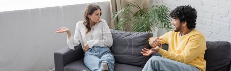 Photo for Emotional brunette woman in white blouse gesturing while talking to young and curly indian man in yellow jumper sitting on sofa near green plant against brick wall in radio studio, banner - Royalty Free Image