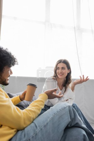 curly indian man in yellow jumper and denim jeans holding coffee to go while gesturing and talking to positive brunette colleague in white blouse on sofa in broadcasting studio