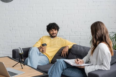 Photo for Brunette woman writing in notebook near curly and smiling indian man in yellow jumper holding coffee to go while talking on couch near microphone and laptop in radio studio - Royalty Free Image