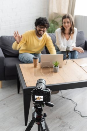 homme indien flou agitant la main pendant le chat vidéo sur ordinateur portable près d'un collègue souriant et appareil photo numérique professionnel en studio de radiodiffusion avec tasse en papier et pot de fleurs sur la table