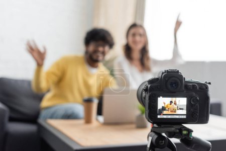 enfoque selectivo de la cámara digital profesional cerca de difuminados vloggers interracial agitando las manos cerca de la computadora portátil y el café para ir mientras se graba podcast en el estudio de radio