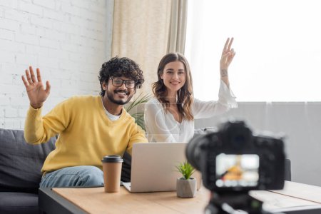 cheerful interracial vloggers in casual clothes waving hand and showing victory gesture during video call on laptop near blurred digital camera in radio studio