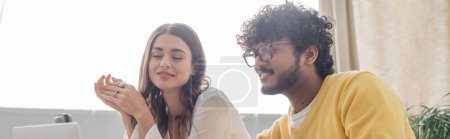 charming brunette woman and curly indian man in eyeglasses and yellow jumper looking at blurred laptop and smiling during video call in podcast studio, banner