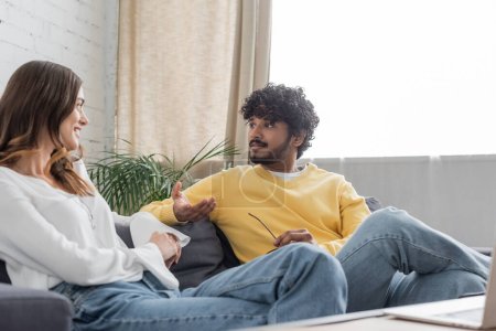 bearded and curly indian radio host in yellow jumper sitting on couch and talking to charming and positive colleague in white blouse while recording podcast in studio