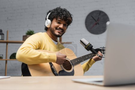 junger, fröhlicher indischer Musiker mit Kopfhörer und gelbem Pullover, der Akustikgitarre spielt, während er im Studio Musik in der Nähe von professionellem Mikrofon und verschwommenem Laptop aufnimmt