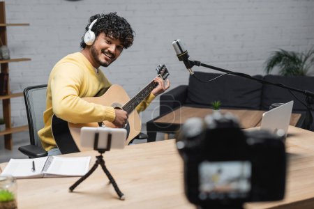 happy indian musician in headphones and yellow jumper playing acoustic guitar near smartphone on tripod, laptop, notebook and digital camera on blurred foreground in radio studio