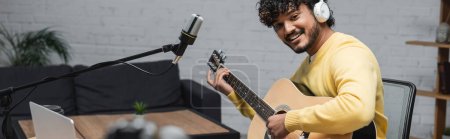 musicien indien bouclé et heureux en casque et pull jaune jouant de la guitare acoustique près d'un ordinateur portable et microphone professionnel en studio avec canapé sur fond, bannière