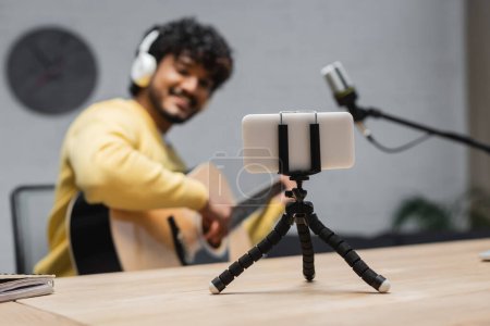 focus on smartphone on tripod near blurred indian musician in yellow jumper and headphones playing acoustic guitar close to professional microphone in broadcasting studio