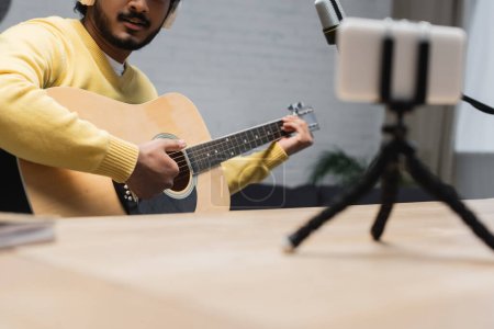 Ausgeschnittene Ansicht eines kreativen indischen Musikers im gelben Pullover, der Akustikgitarre in der Nähe von Mikrofon und Smartphone mit Stativ spielt, während er Podcast auf verschwommenem Vordergrund aufnimmt