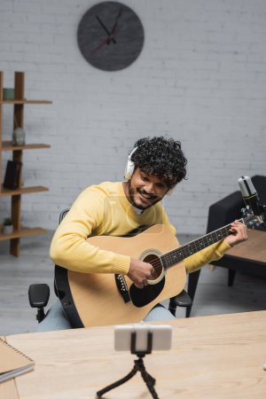 fröhlicher indischer Musiker mit Kopfhörer und gelbem Pullover spielt Akustikgitarre in der Nähe von verschwommenem Handy auf Stativ und professionellem Mikrofon, während er Podcast im Studio aufnimmt