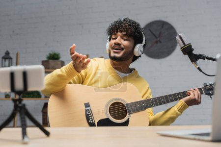 happy and curly indian podcaster in headphones and yellow jumper holding acoustic guitar and pointing with hand near blurred laptop and smartphone on tripod in record studio