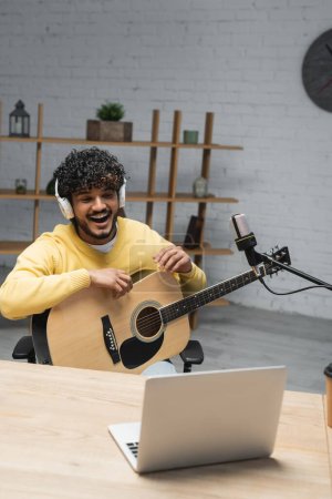 hombre indio rizado en auriculares inalámbricos y puente amarillo riendo mientras se sienta con la guitarra acústica cerca de la computadora portátil y micrófonos profesionales en el estudio