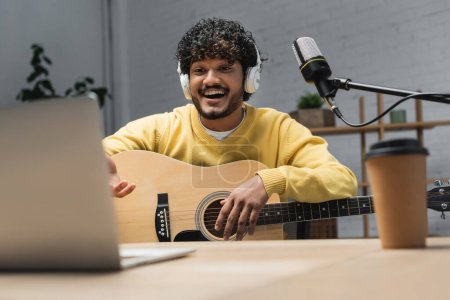fröhlicher indischer Musiker mit Kopfhörer und gelbem Pullover spielt Akustikgitarre in der Nähe von verschwommenem Laptop und Studiomikrofon auf Tisch mit verschwommenem Coffee to go während der Aufnahme eines Podcasts im Studio