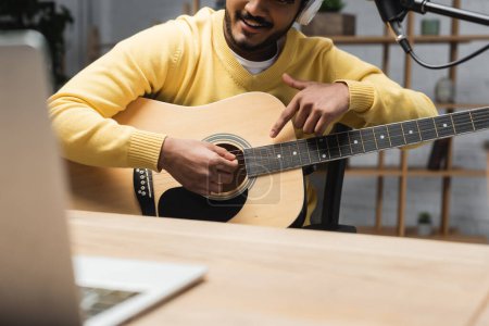 vista recortada de hombre indio sonriente en puente amarillo apuntando con el dedo a la guitarra acústica cerca de la computadora portátil borrosa en la mesa de madera en el estudio de radiodifusión