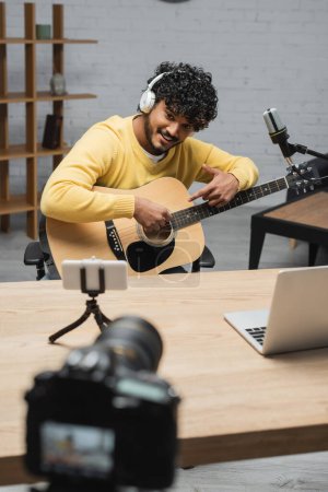smiling indian musician in headphones pointing at acoustic guitar while sitting near laptop and smartphone on tripod in front of blurred digital camera in professional studio