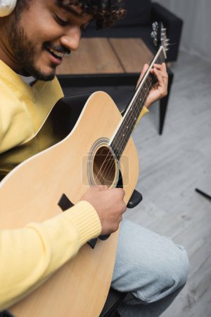 Blurred and smiling young indian musician in casual clothes and wireless headphones singing and playing acoustic guitar during performance in studio  puzzle 653051034