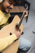Blurred and smiling young indian musician in casual clothes and wireless headphones singing and playing acoustic guitar during performance in studio  Poster #653051034