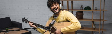 Wesoły młody indyjski podcaster w słuchawkach bezprzewodowych gra na gitarze akustycznej w pobliżu mikrofonu podczas strumienia w studio podcast, baner 