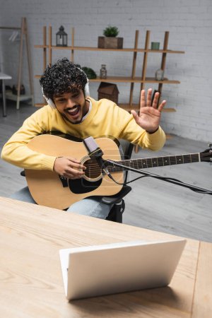 Téléchargez les photos : Podcaster indien joyeux dans les écouteurs tenant la guitare acoustique et agitant la main à l'ordinateur portable sur une table en bois près du microphone pendant le flux en studio - en image libre de droit
