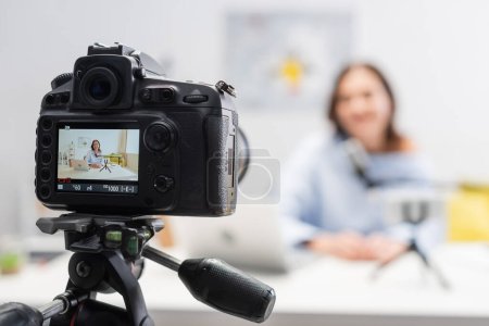 Digital camera on tripod standing near blurred happy female podcaster using devices and microphone on wooden table during stream in podcast studio 