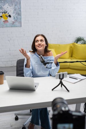 Smiling brunette podcaster looking away while talking near microphone, devices, coffee to go and notebook on wooden table during stream in studio 