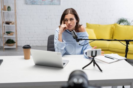 Pensive brunette broadcaster looking away while talking near microphone, smartphone on tripod, laptop and coffee to go on table during stream in podcast studio 