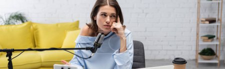 Pensive brunette blogger looking away while talking near microphone, mobile phone and coffee to go during stream in podcast studio, banner 