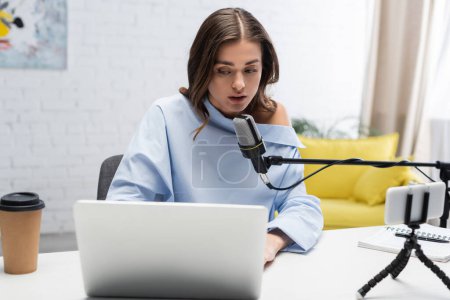 Brunetka nadawca w bluzce rozmawia blisko mikrofonu i za pomocą laptopa w pobliżu kawy, aby przejść, notebook, długopis i smartfon podczas strumienia w studio 