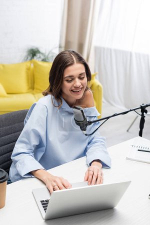 Carefree brunette broadcaster using laptop near microphone during stream, near coffee to go and notebook on table while sitting in podcast studio 