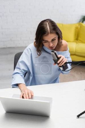 Brünette Moderatorin in blauer Bluse, die am Mikrofon spricht und einen verschwommenen Laptop benutzt, während sie beim Online-Stream im Podcast-Studio neben dem Tisch sitzt, mit gelbem Sofa auf verschwommenem Hintergrund 