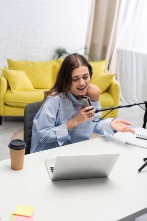 Pozytywne i brunetka podcaster rozmowy na mikrofon, gestykulując i patrząc na laptopa w pobliżu kawy, aby przejść i notebook na stole podczas strumienia w studio 